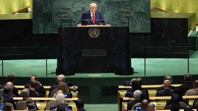 O presidente dos EUA, Donald Trump, vestido com terno azul-escuro, camisa branca e gravata vermelha, discursa na Assembleia Geral da ONU, em Nova York, a partir de um púlpito com o brasão da ONU, enquanto os membros estão sentados em suas mesas, sobre um palco verde