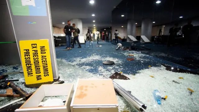 Detalhe do Congresso Nacional com vidros quebrados no chão e um cartaz: 